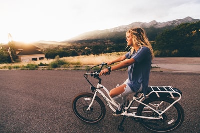 白天，一位身穿蓝色t恤和破旧的蓝色牛仔牛仔裤的妇女骑着白色自行车在山区附近的灰色道路上行驶